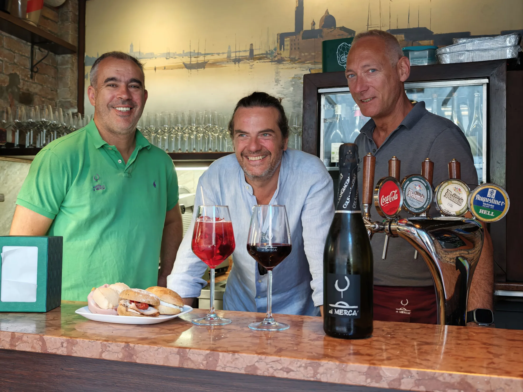 The owners of Al Mercà at the bacaro counter with a guest, glasses of wine and a cicchetto, the Venice lagoon in the background
