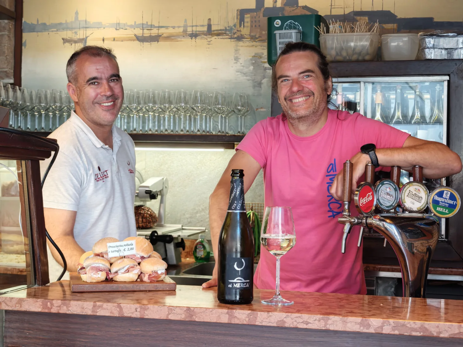The two smiling owners of Al Mercà behind the bacaro counter at Rialto, with a board of cicchetti and a bottle of white wine